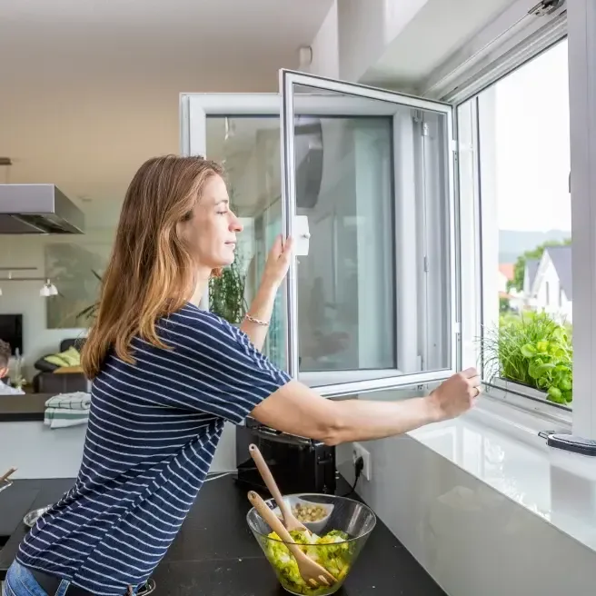 Frau öffnet in der Küche ein Fenster mit Fliegengitter-Drehrahmen